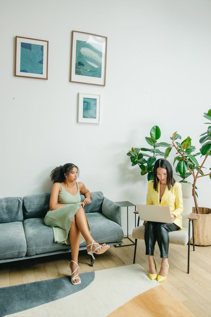 Two women in a stylish living room setting, one using a laptop and the other relaxing.